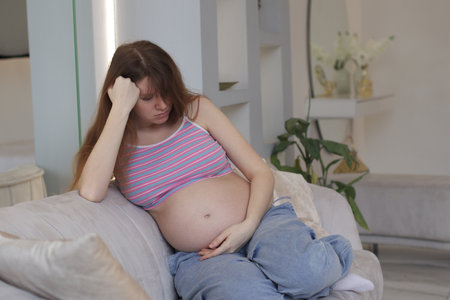 Young woman feel depression suffering from nausea or belly ache and headache during pregnancy. Sitting in a sofa in the living room at home, looking unhappy and worriedの写真素材