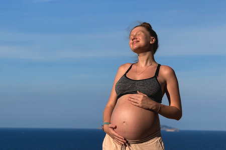 Young pregnant woman stroking her belly outdoors on sea, beach at summer vacation