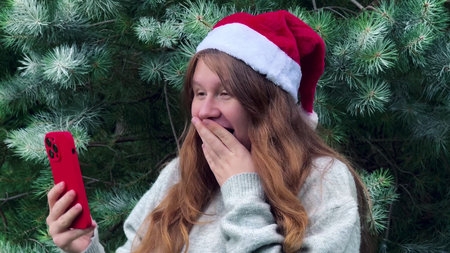 Happy positive young woman in Christmas hat using her cell mobile phone outdoors, wishing Merry Christmasの写真素材