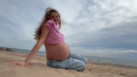 beautiful pregnant woman with big belly and long hair sits on sand by sea, water and enjoys weatherの写真素材