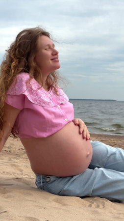 beautiful pregnant woman with big belly and long hair sits on sand by sea, water and enjoys weatherの写真素材