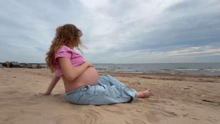 beautiful pregnant woman with big belly and long hair sits on sand by sea, water and enjoys weatherの写真素材