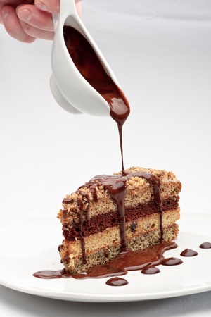 A piece of fresh baked delicious sponge-cake with chocolate sauce on a white plate. A hand pouring chocolate sauce out of a sauceboat on it.の写真素材