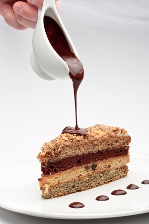 A piece of fresh baked delicious sponge-cake with chocolate sauce on a white plate. A hand pouring chocolate sauce out of a sauceboat on it.の写真素材
