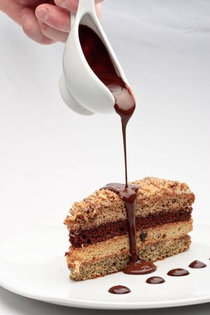 A piece of fresh baked delicious sponge-cake with chocolate sauce on a white plate. A hand pouring chocolate sauce out of a sauceboat on it.の写真素材