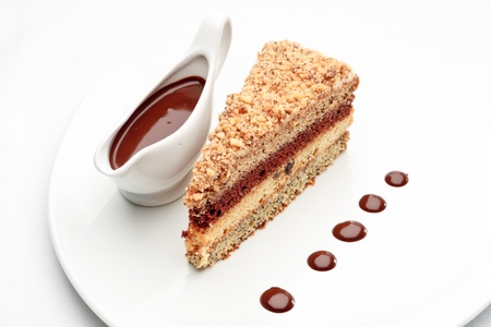 A piece of fresh baked delicious sponge-cake with chocolate sauce on a white plate. A hand pouring chocolate sauce out of a sauceboat on it.の写真素材