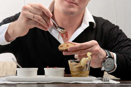 A young man spreading cherry jam on a piece of bread, sitting at an elegantly served table in a restaurantの写真素材