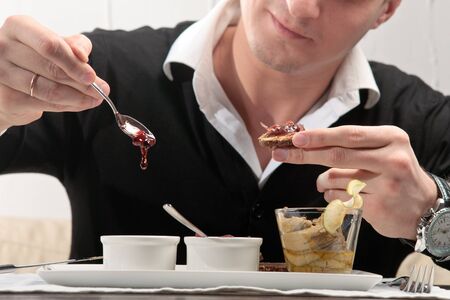 A young man spreading cherry jam on a piece of bread, sitting at an elegantly served table in a...
