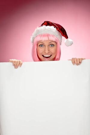 Young female santa in pink wig holding a christmas clear white card over red backgroundの写真素材
