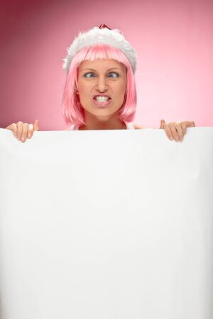 Young female santa in flaunting pink wig holding a christmas clear white card over red backgroundの写真素材