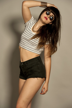 Beautiful young retro female brunette with straight hair in a studio setting while wearing a short top and black shorts with sunglasses on a gray background.の写真素材
