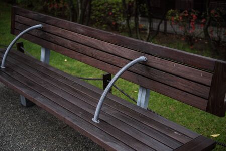 A sitting bench made of wood is seen on the picture inside a park. On the background, the lush...