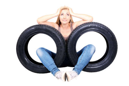 Beautiful young woman sitting between two tyres and thinking of what she should do. Isolated on white background.の写真素材