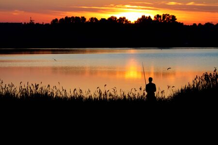 Fisherman fishing at a lake at sunset. Backlit silhouette photoの写真素材