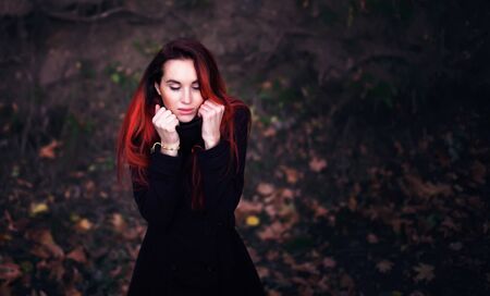 Beautiful woman with dyed red hair in a warm coat bundling up to keep herself warm during cold autumn weatherの写真素材