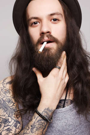 The guy is smoking and looking.handsome bearded man in the hat smoking a cigarette.Brutal hipster boy with tattoo and long hairの写真素材
