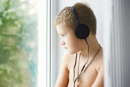 little boy loking in window and listening music in headphones. lifestyle portrait of child at homeの写真素材