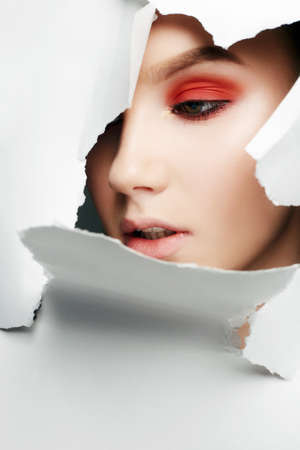 Face of a young beautiful girl with a bright make-up into a hole in paper. Beauty makeup, orange eyes shadowsの写真素材