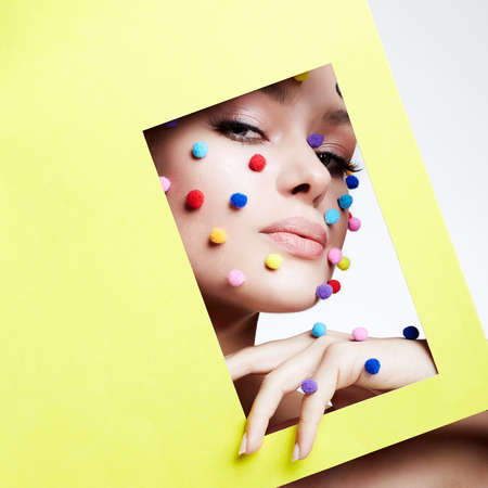 Beautiful young woman into the hole of color paper. Beauty portrait of  girl with colorful balls on her face. make-up conceptの写真素材