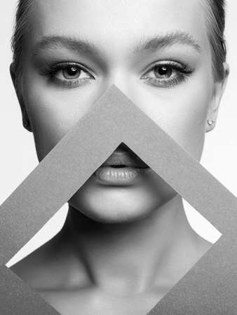 Beautiful young woman Face in paper triangle. Beauty black and white portrait of girl with make-upの写真素材
