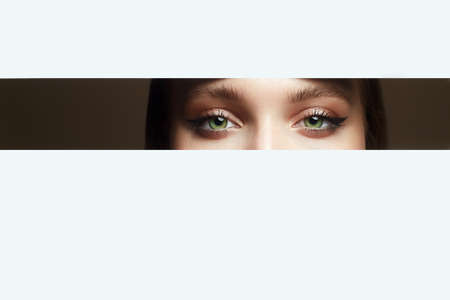 beautiful eyes peep into the crack. young beautiful woman. make-up artist concept. A girl with beautiful bright green eyes with shining shadows, looks into the hole of white paperの写真素材