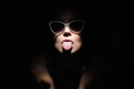 Beautiful woman in glasses, showing tongue. Retro style portrait. Lady Girl looking from the dark