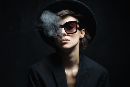 Beautiful woman in sunglasses and hat. Lady in suit releasing smoke from mouthの写真素材