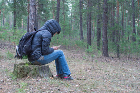 A woman sits on a stump in the woods early in the morning and checks her cellphone.  A female hiker, backpacker trekking in the woods and mountains. Healthy lifestyle adventure, camping on hiking trip.の写真素材