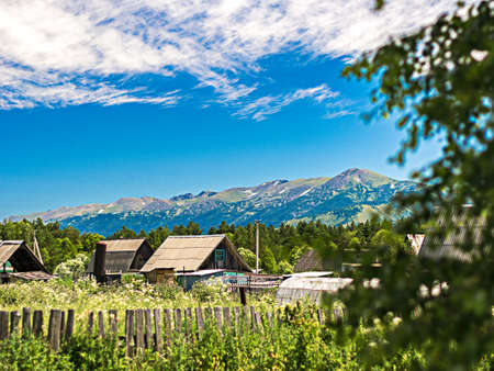 Roofs of traditional houses seen behind the fence and gardens in Altai Mountains village, Kazakhstan, with a ridge in background on a sunny summer dayの写真素材