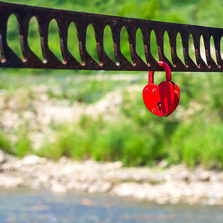 A single heart shaped red padlock symbolizing love hanging on the bridge handrails with blurred river, greenery, hill in backgroundの写真素材