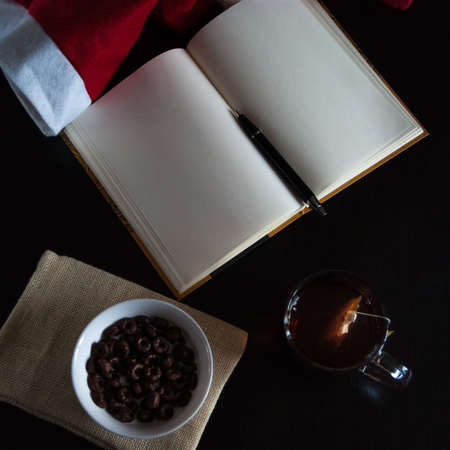 Planning future on Christmas: a notebook with blank pages, a black pen, Santa hat, glass mug with teabag, white bowl of chocolate cereal rings and milk on plain cloth, all on dark dining tableの写真素材