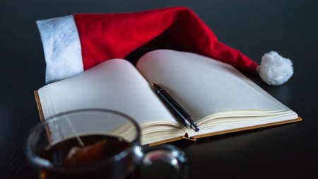 Planning future on Christmas concept: A notebook with blank pages, a black pen, Santa hat, glass mug with teabag, all on dark dining tableの写真素材