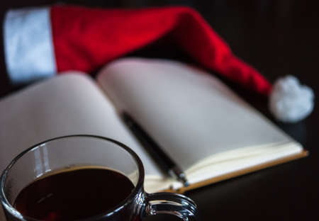 Planning future on Christmas concept: a notebook with blank pages, a black pen, Santa hat, glass mug with teabag, all on dark dining tableの写真素材