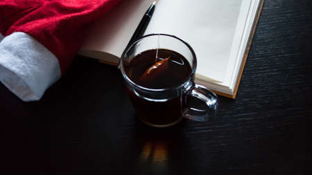 Planning future on Christmas concept: A notebook with blank pages, a black pen, Santa hat, glass mug with teabag, all on dark dining tableの写真素材