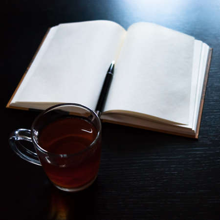 A glass mug with tea and a lemon slice, notebook with blank pages, black pen on dark deskの写真素材