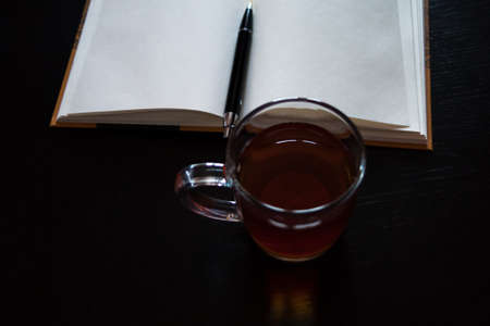 A glass mug with tea and a lemon slice, notebook with blank pages, black pen on dark deskの写真素材