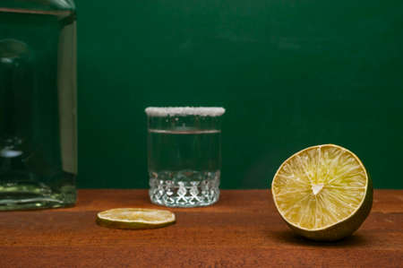 Half Of A Lime With Blurred Lime Slice, Silver Tequila Shot Rimmed With Salt And A Bottle On A Wooden Rustic Table. Green Background.の写真素材