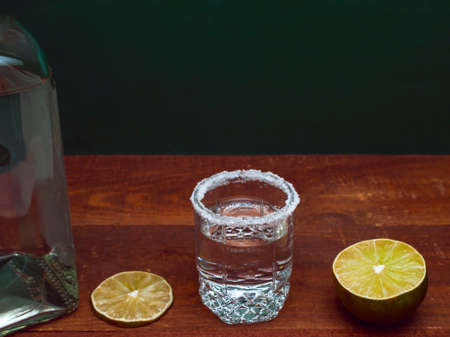 Alcohol Silver Tequila Shot Rimmed With Salt On A Wooden Rustic Table. Cross Section Of A Lime, A Lime Slice And A Bottle. Green Background.の写真素材