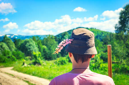 Back of a Guy in a Pink T-Shirt, Bucket Hat with Raceme of Pink Fireweed, Chamaenerion, and a Pole on a Mountain Road, Woodland Background on a Summer Day.の写真素材
