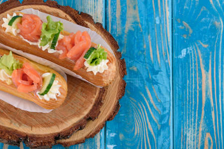 Tasty and beautiful eclairs on a wooden board. Appetizing dessert with red fish, cucumber, cheese and greens. With blue wood .の写真素材