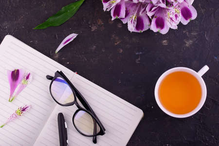 View from above. Fragrant tea in a white cup, beautiful flowers, a black background, a notebook with a pen and glasses. Office.の写真素材