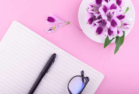 View from above. Fragrant tea in a white cup, beautiful flowers, a pink background, a notebook with...