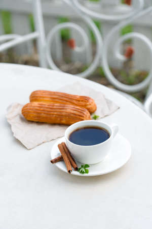 Fragrant coffee in a white cup with eclairs in a summer cafe. Spring, cinnamon, leaf sprigs.