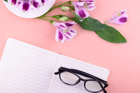 View from above. Fragrant tea in a white cup, beautiful flowers, a pink background, a notebook with a pen and glasses. Office.の写真素材