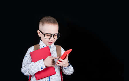 A boy with a backpack with glasses holds a red notebook and red phones on a black background. School...