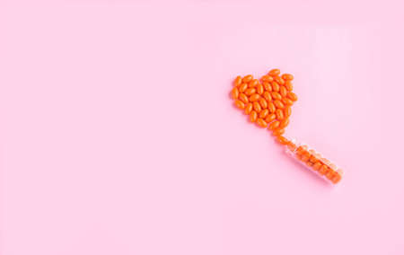 Orange tablets are poured out of a glass flask in the shape of a heart on a pink background.