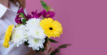 The girl on a pink background holds a beautiful bouquet of different flowers and covers her face....