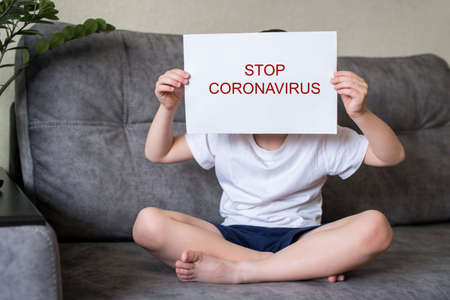 The boy sits on the couch holding a white sheet of paper in front of his face with the inscription stop coronavirus, and covers his face. Home quarantineの写真素材