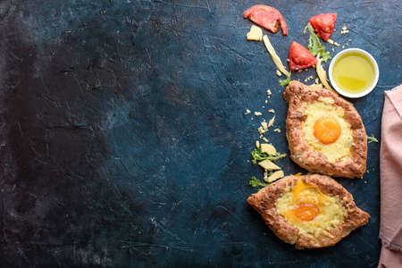 Two fresh and delicious khachapuri of weed grain flour and cottage cheese, egg, sulugini cheese, vegetables, oil, cutlery on a dark background.の写真素材