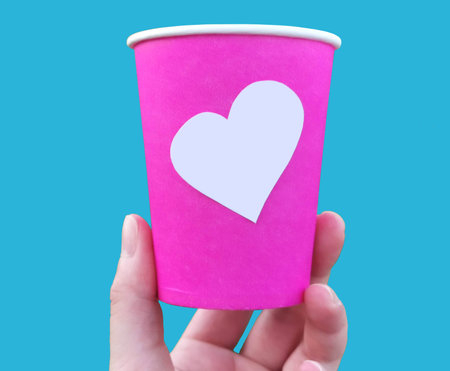 Holding pink coffee cup with blank heart shape sticker. Hand holds paper cup image isolated on plain background. Empty heart shaped place for you designの写真素材